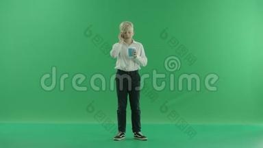 一个穿着<strong>校服</strong>的金发小男孩正拿着一杯咖啡或茶，用手机说话。 他站着